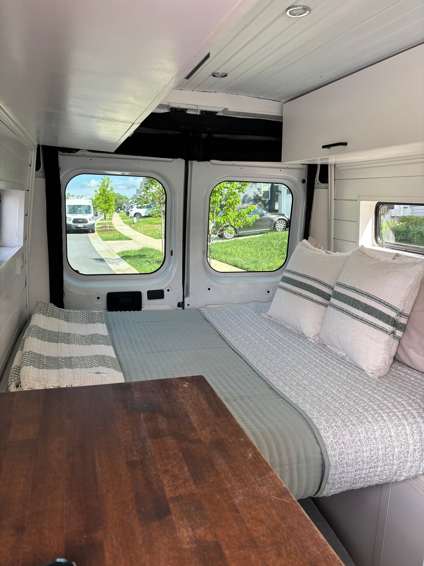 Cozy RV rental interior: bed with pillows, wooden table, and street views through large windows, perfect for travel.