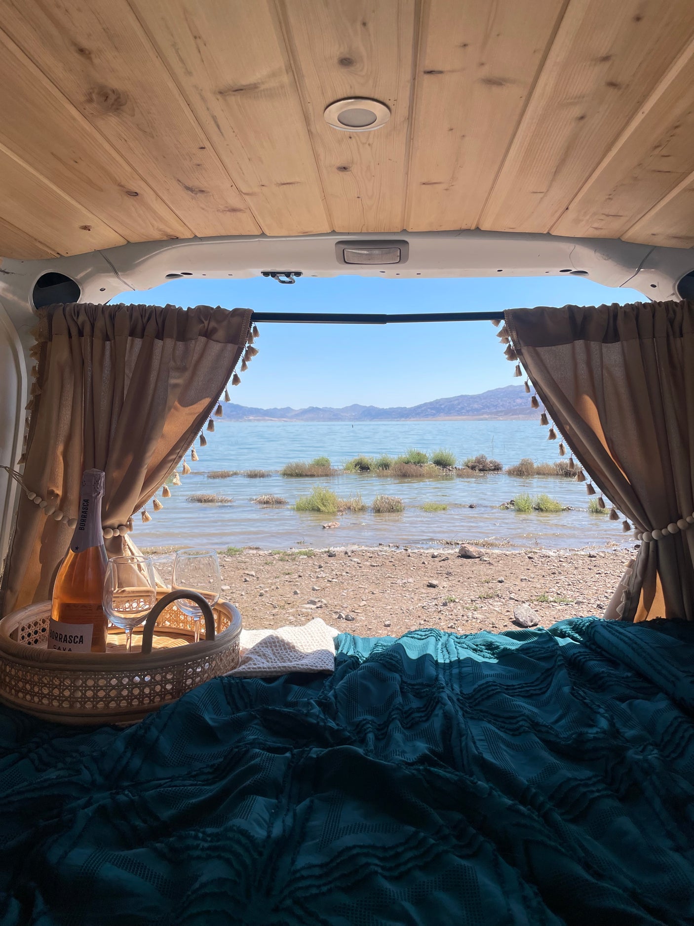 Enjoying a serene lake view from an RV rental, featuring a cozy bed, pulled-back curtains, and sparkling wine on a rattan tray.