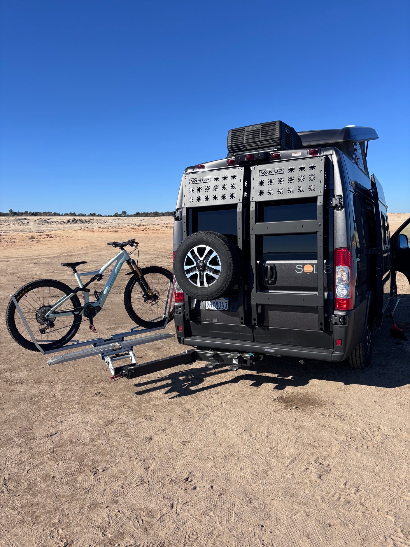 RV rental: Dark grey camper van with roof AC & bike rack, holding a light mountain bike, parked in sandy terrain.