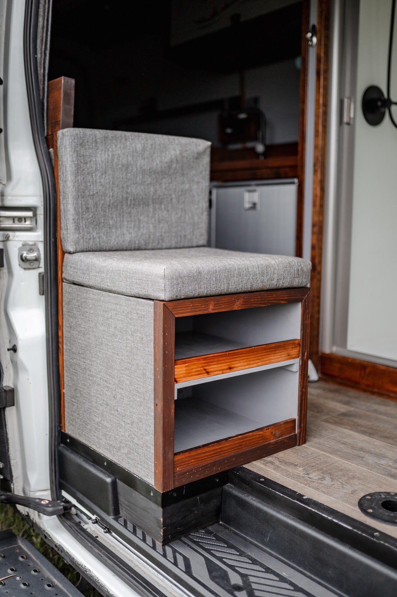 Cozy RV rental interior: grey upholstered seat with custom wood storage cubbies & integrated fridge for practical camper van living.