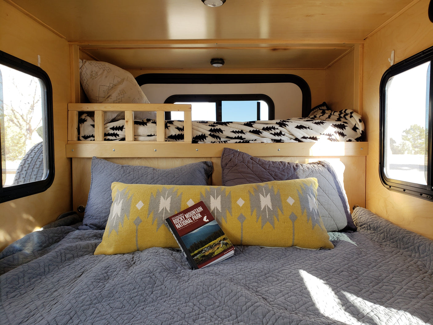 Cozy RV rental interior shows a lower bed with a national park guide, patterned pillows, and a raised bunk bed. Perfect for trips!