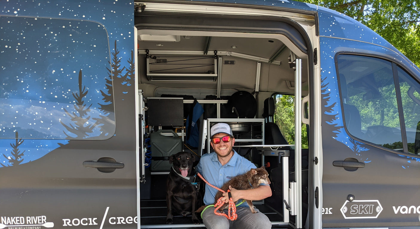 A man and his two dogs pose inside a spacious RV rental van with custom starry night wrap, ideal for outdoor trips.