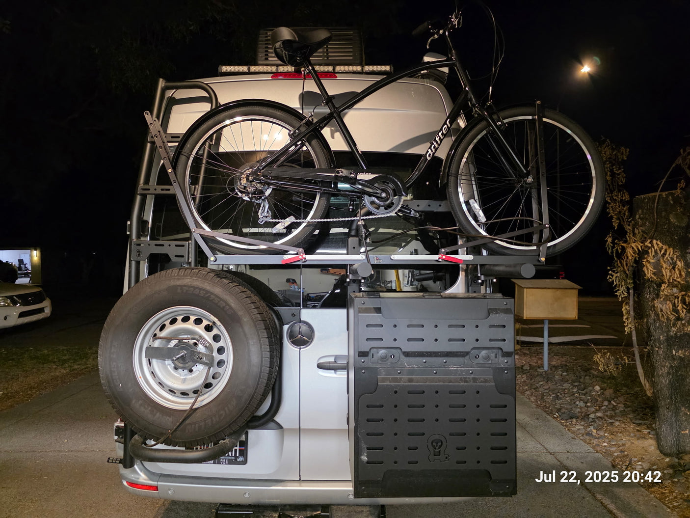 RV rental: A silver Mercedes van's rear shows a bike, spare tire, & side storage. Night scene, ready for adventure.