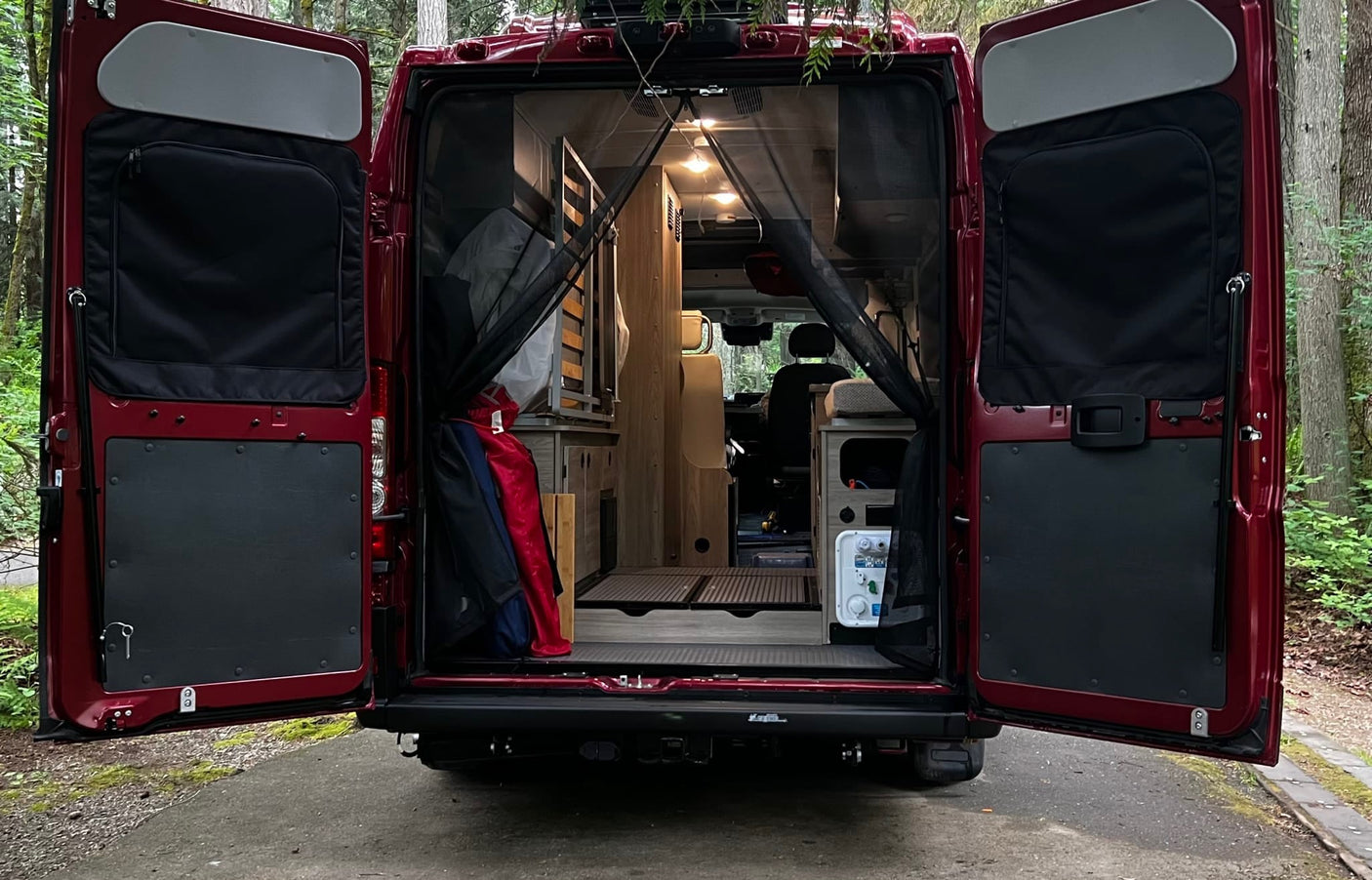 Rear view of a red RV rental with open doors, revealing a custom-built interior with bed platform & storage, parked in a forest.