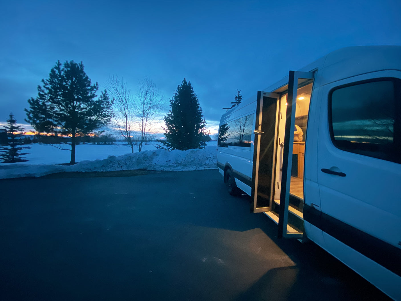 Experience winter travel with an RV rental! A white camper van glows from within at dusk, surrounded by snowy fields and trees.