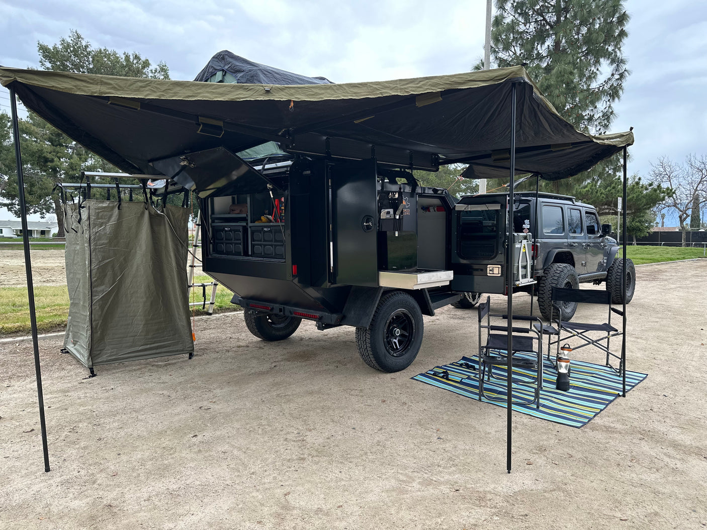 Full overlanding camp setup: A black expedition trailer & Jeep with an extended awning, outdoor kitchen & shower tent. Ideal for rv rental.