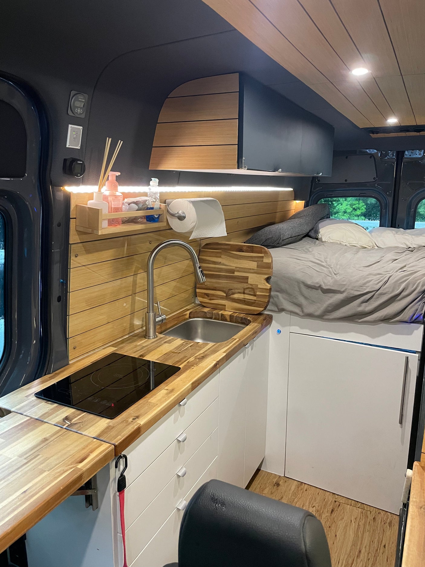 RV rental interior: modern kitchen with wood counters, induction cooktop, sink, white cabinets, and a comfortable bed.