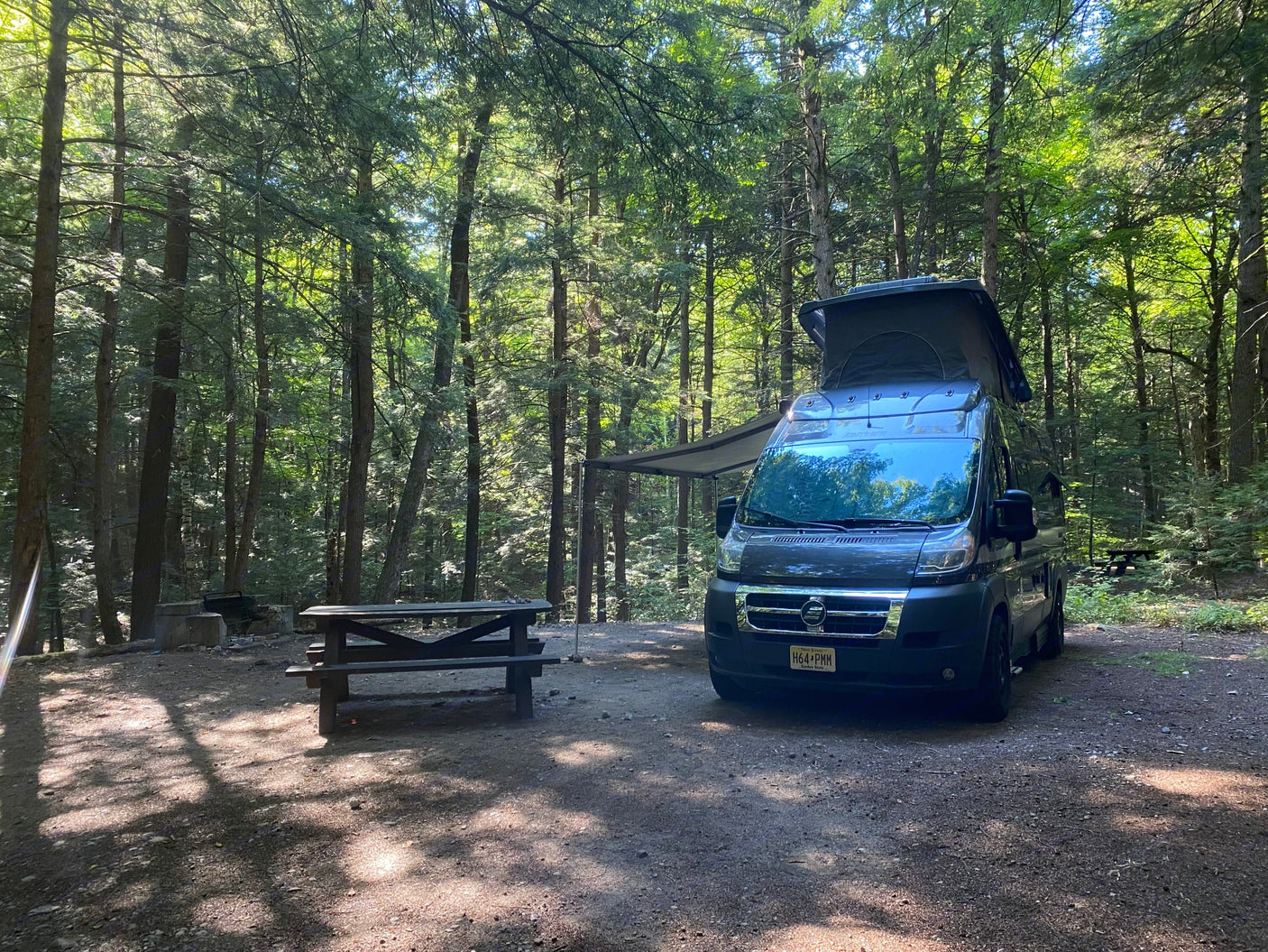 An RV rental camper van with a pop-top and awning set up at a forest campsite. A picnic table and fire pit complete the outdoor scene.