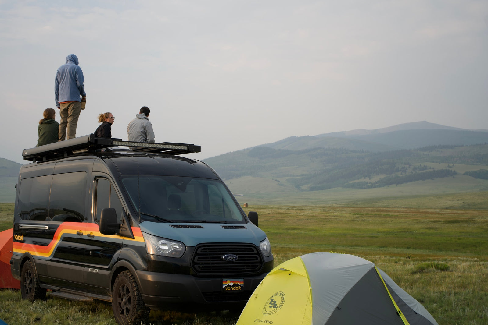 Vandoit RV rental adventure: People on a customized Ford Transit van roof enjoy vast scenic views and their campsite with tents.