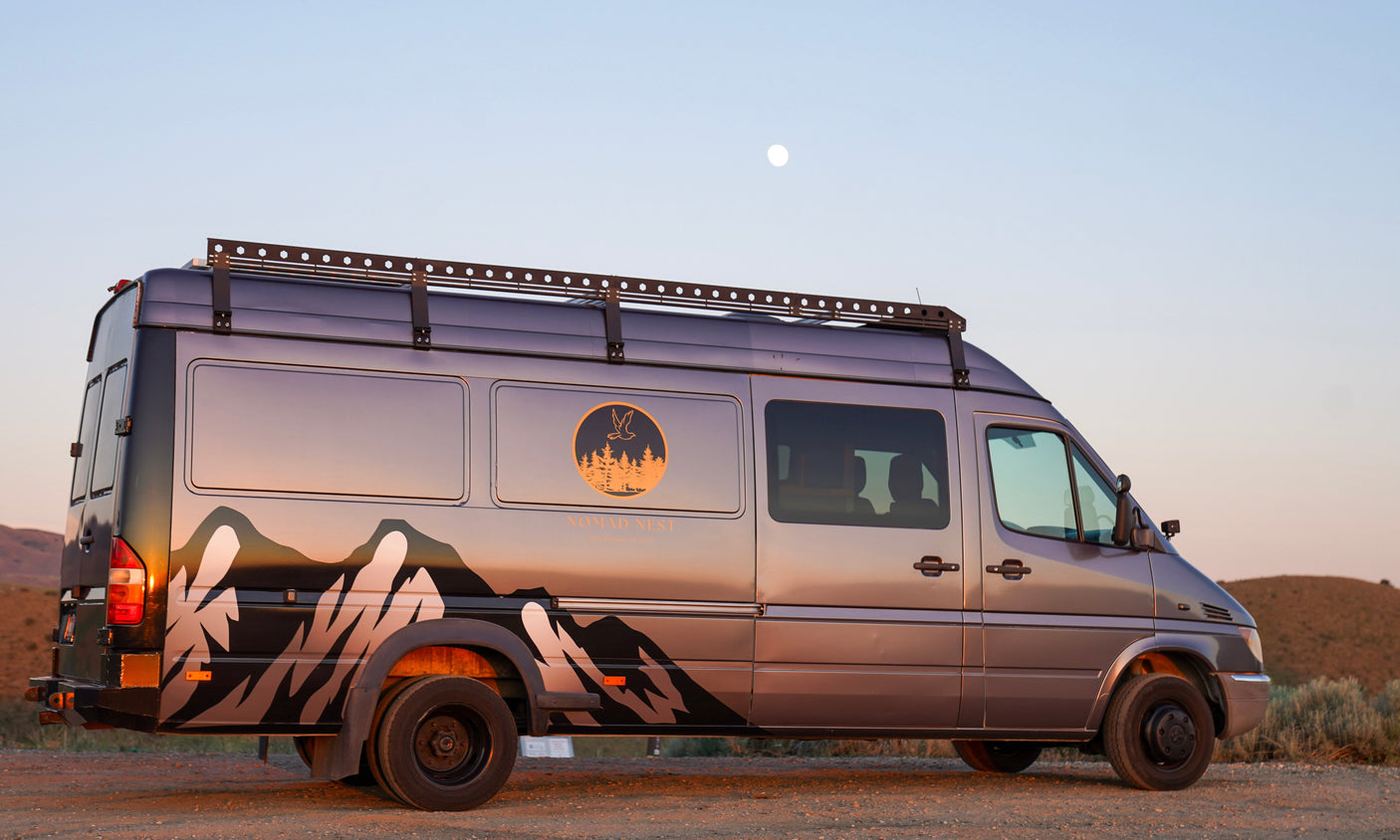 This grey camper van, ideal for RV rental adventures, features mountain graphics and a roof rack under a soft, moonlit sky.