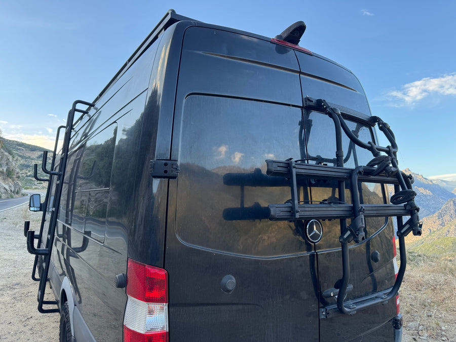 Dark Mercedes-Benz Sprinter RV rental with rear bike rack and side ladder, ready for adventure in the mountains.