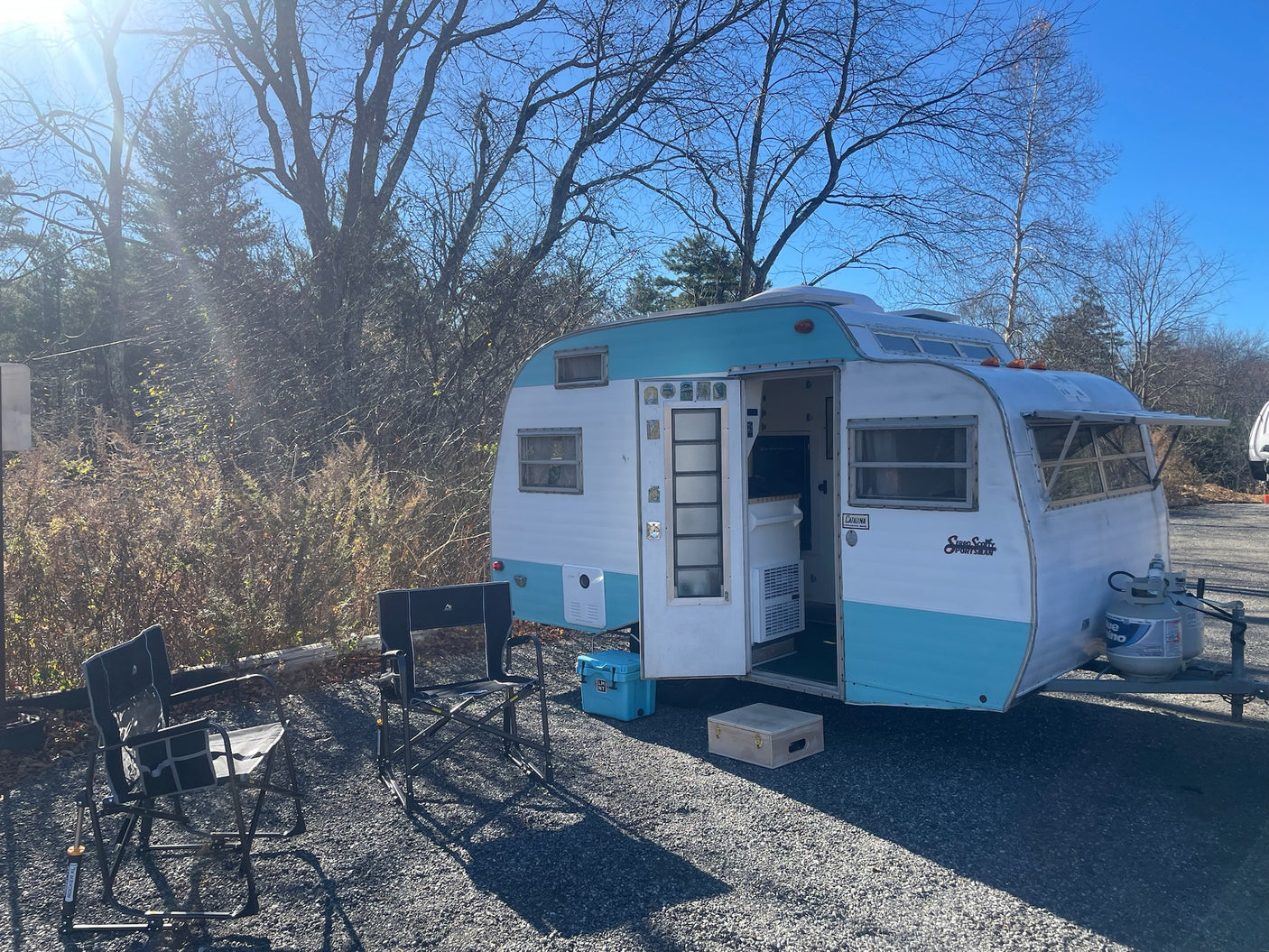 Vintage teal and white travel trailer RV rental, parked on gravel with two chairs. Sunny outdoor scene with bare trees.