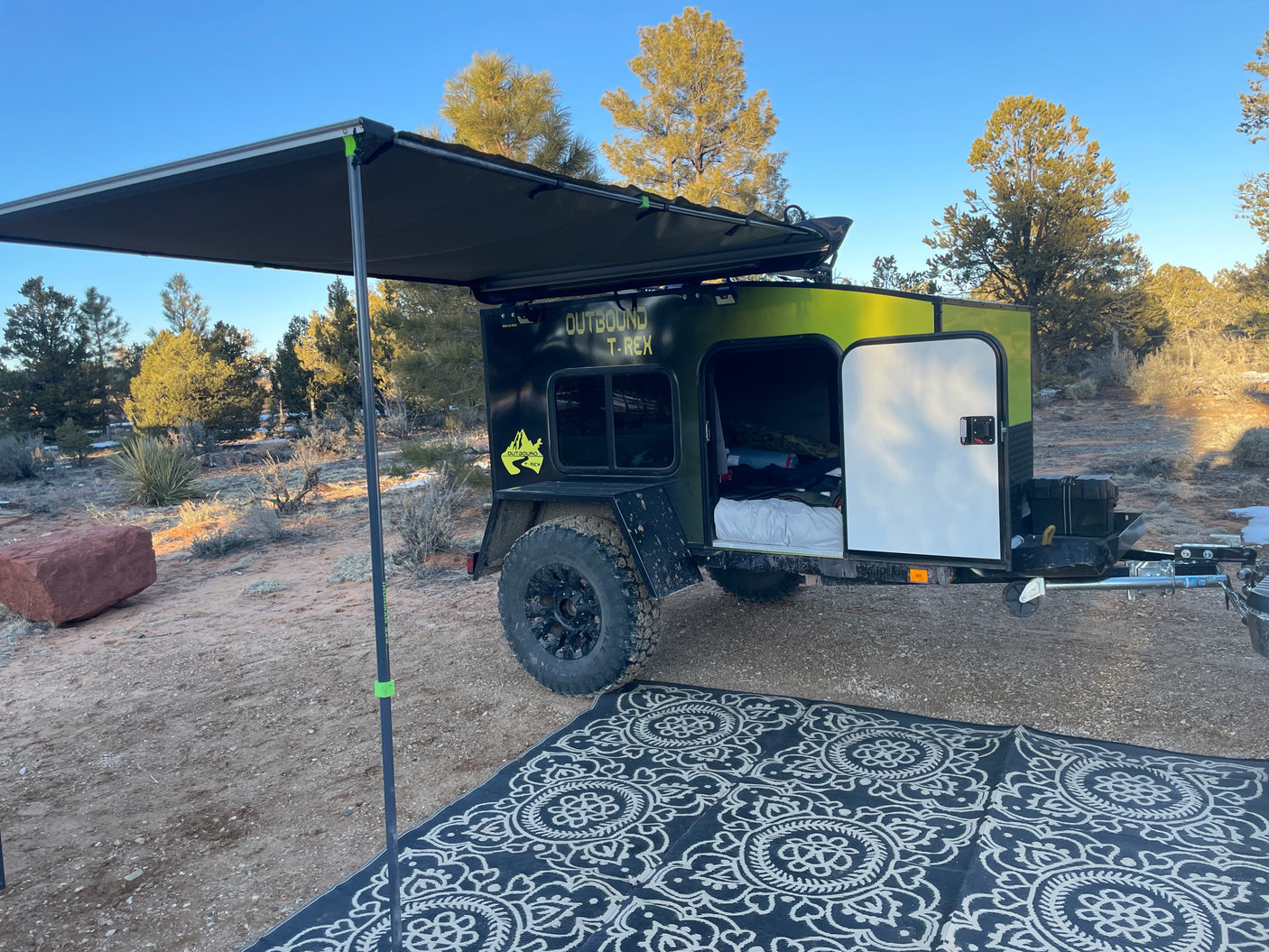 An Outbound T-Rex teardrop camper with awning and rug, perfect for your next RV rental adventure in nature.