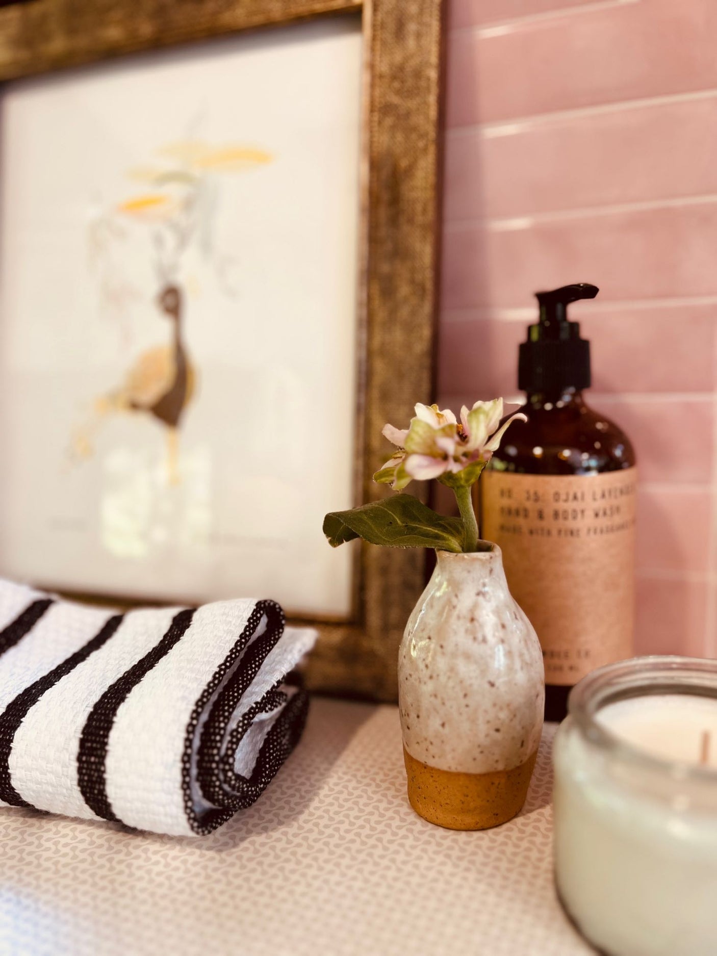Cozy RV rental bathroom: fresh flower in small vase, striped towel, soap, and candle on counter, pink tiled wall.
