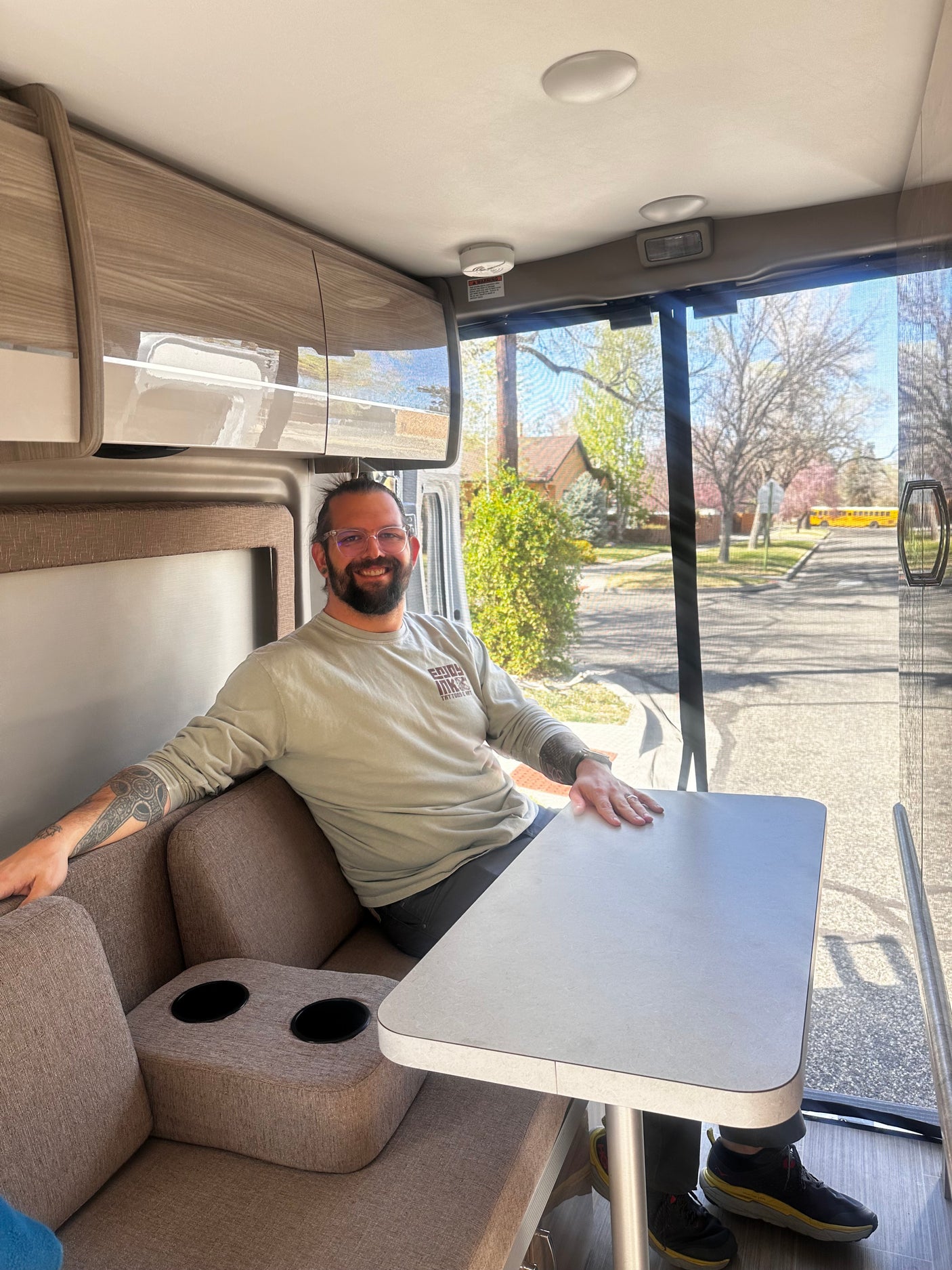 A man smiles from a cozy seat inside an RV rental, with a dining table and large window showing a sunny street.
