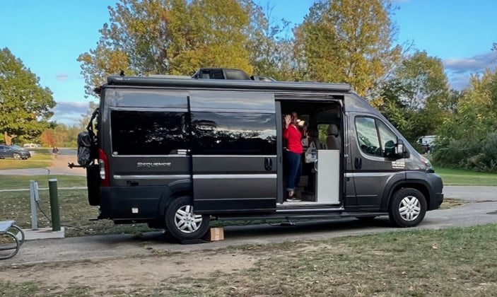 A gray Thor Sequence RV rental camper van parked at a fall campsite. A person waves from inside the open side door.|A happy couple smiles inside their RV rental