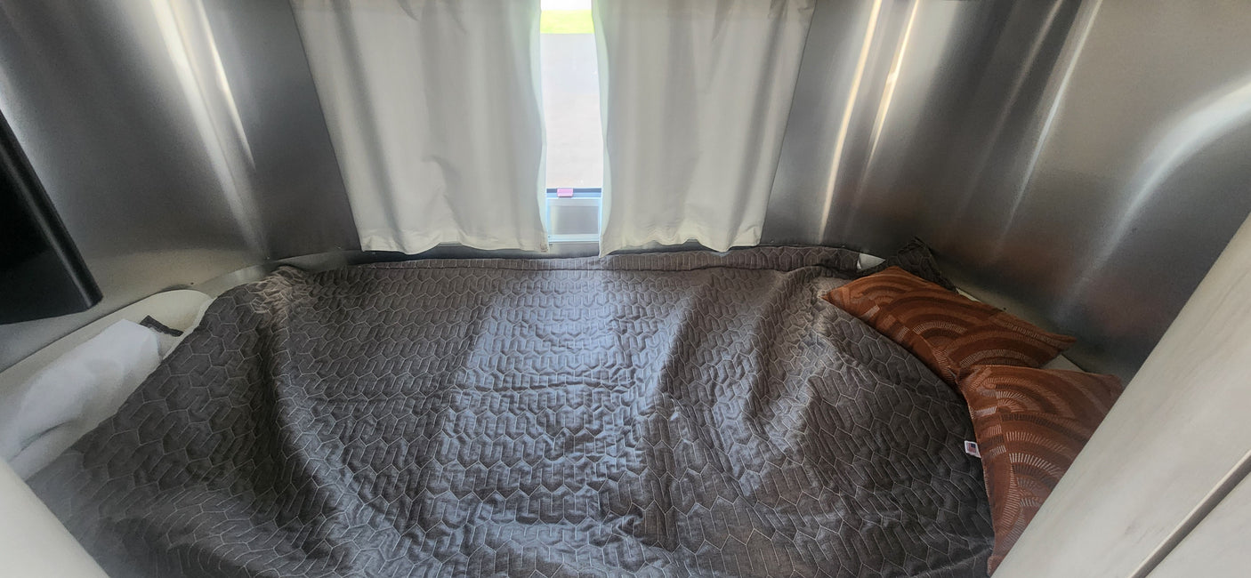 Inside a modern RV rental: a comfortable bed with a gray hexagonal quilt, orange pillows, and metallic walls for cozy travel.
