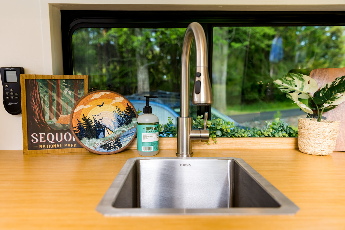 RV rental's inviting kitchen sink area, featuring a scenic window view, wooden counter, and nature-themed decor like an embroidered hoop.
