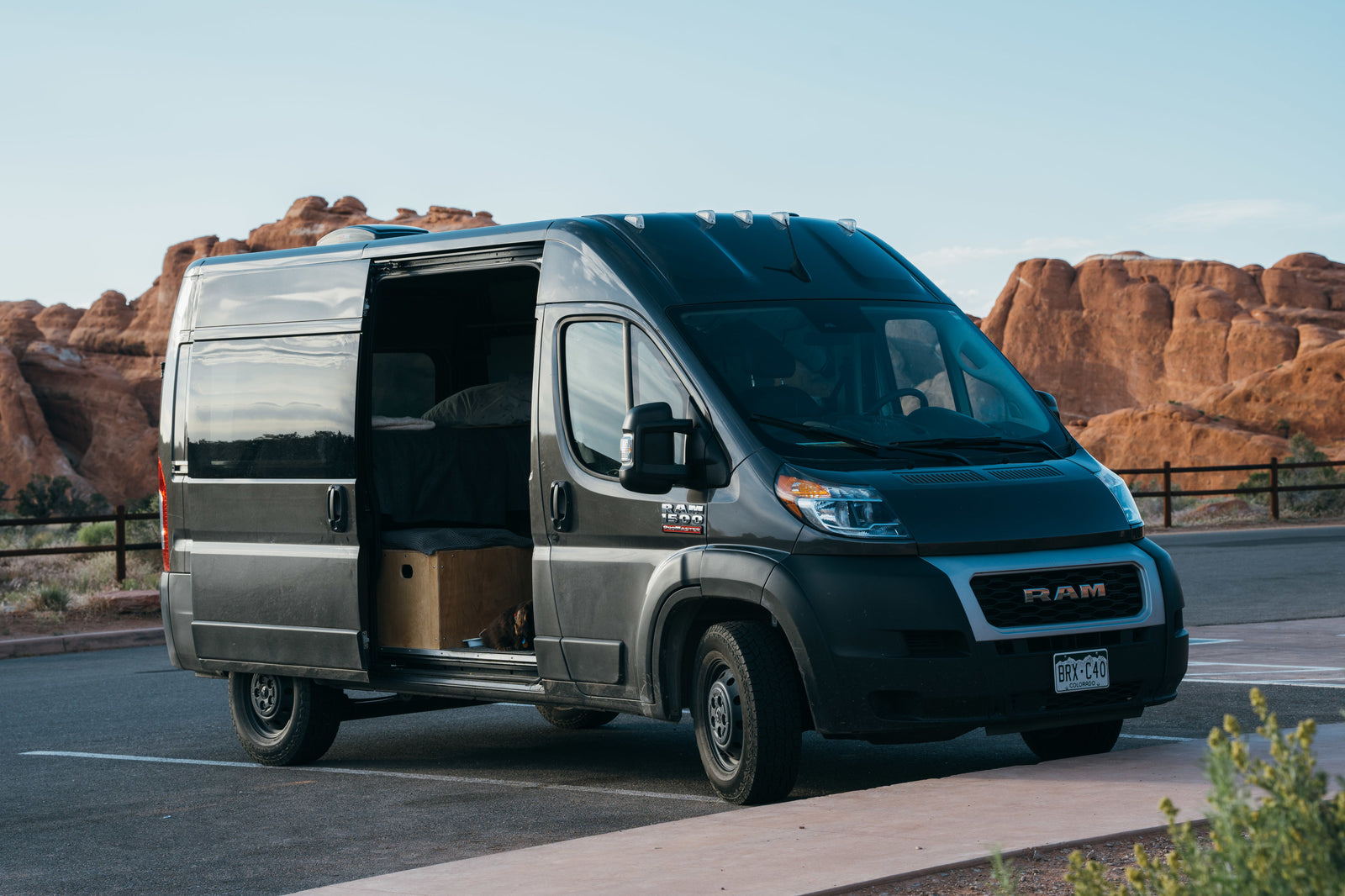 Camper van RV rental: A dark gray Ram ProMaster with its side door open, revealing a bed and dog, parked in a desert landscape.