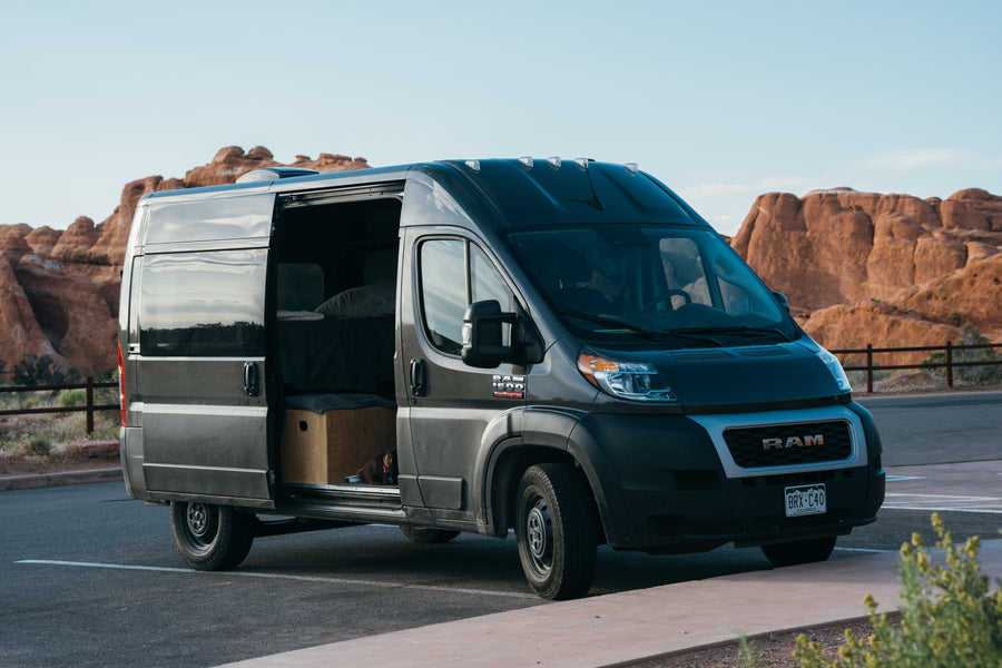 Camper van RV rental: A dark gray Ram ProMaster with its side door open, revealing a bed and dog, parked in a desert landscape.