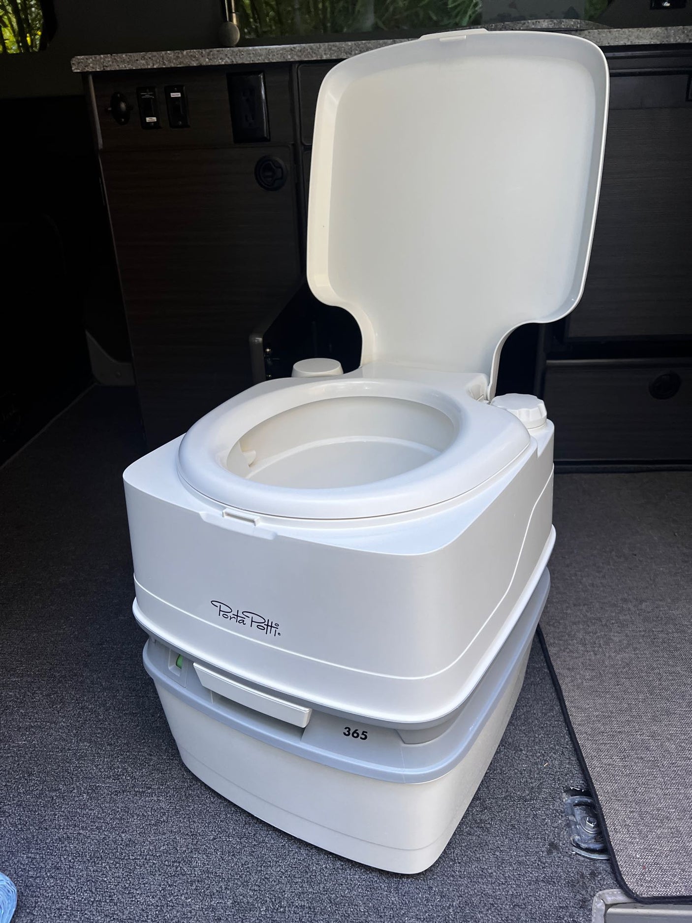 Portable Porta Potti toilet in an RV rental, essential for camping and travel. The white unit sits on dark carpet near wood cabinets.