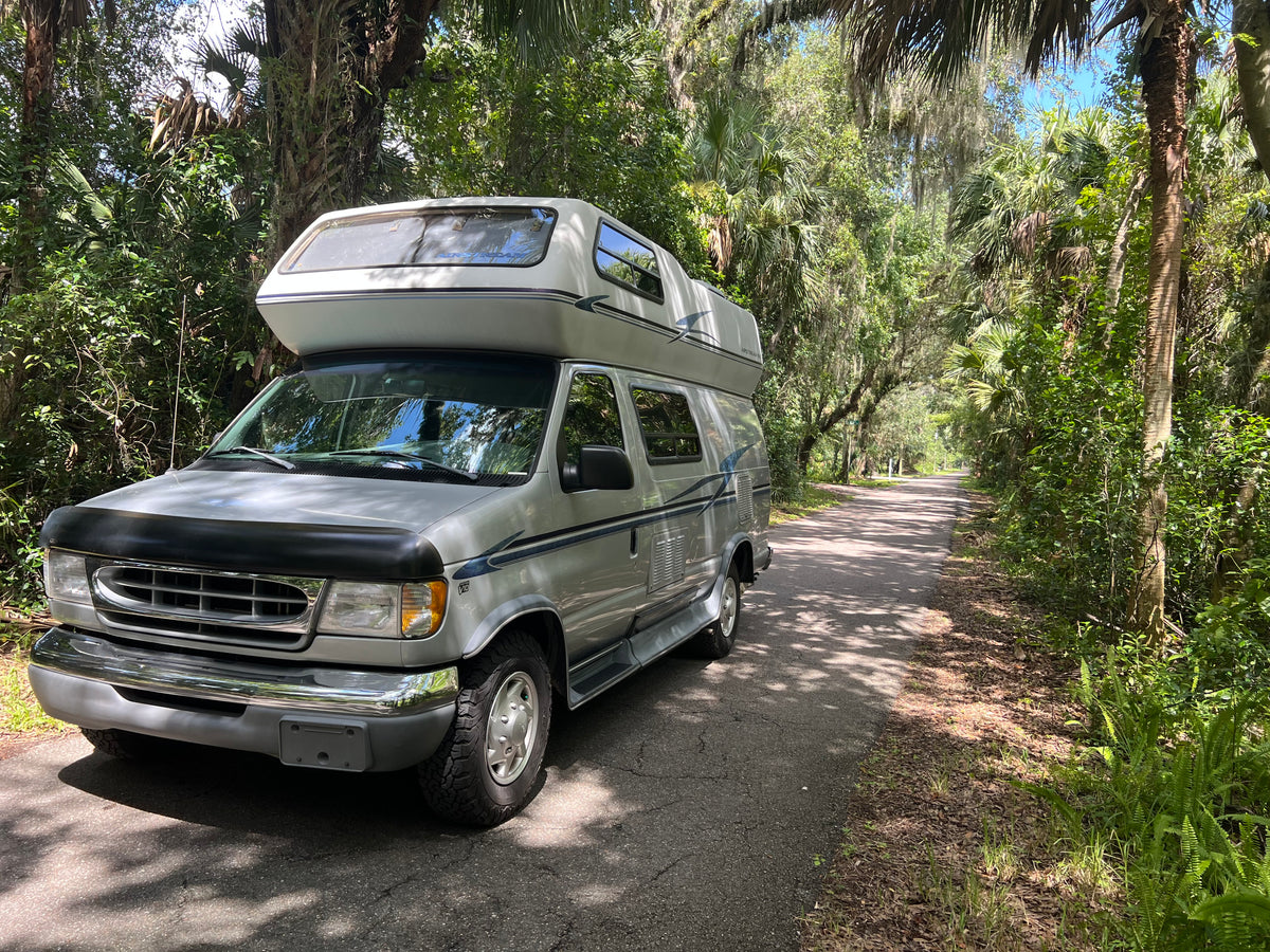 Silver Airstream Ford Econoline camper van rv rental on a shaded road, surrounded by lush, mossy trees and tropical foliage. — photo 7