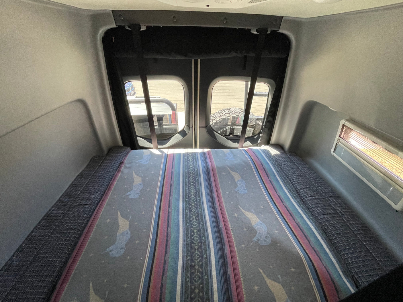 View of a comfortable RV rental bed with a patterned blanket, looking out the rear windows of the vehicle.