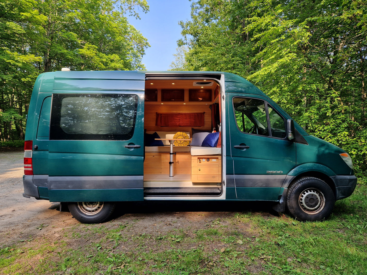Green RV rental van parked in a forest, showcasing its custom wood interior through the open side door.