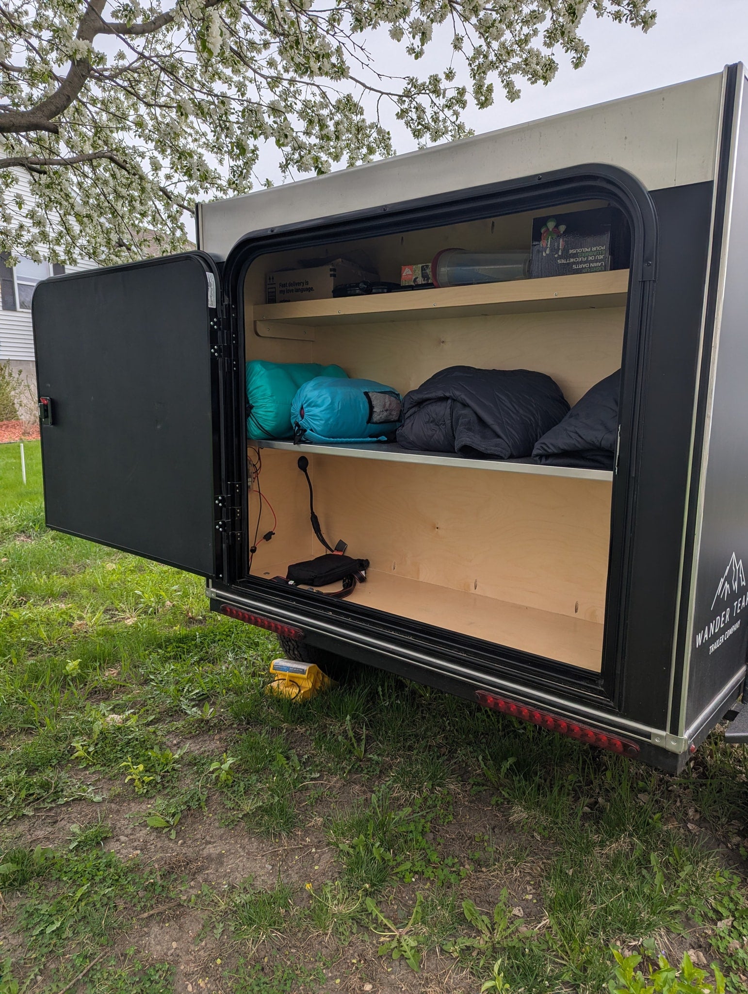 Open side hatch of a Wander Tear RV rental trailer showing organized shelves with sleeping bags & gear, under a blossoming tree.