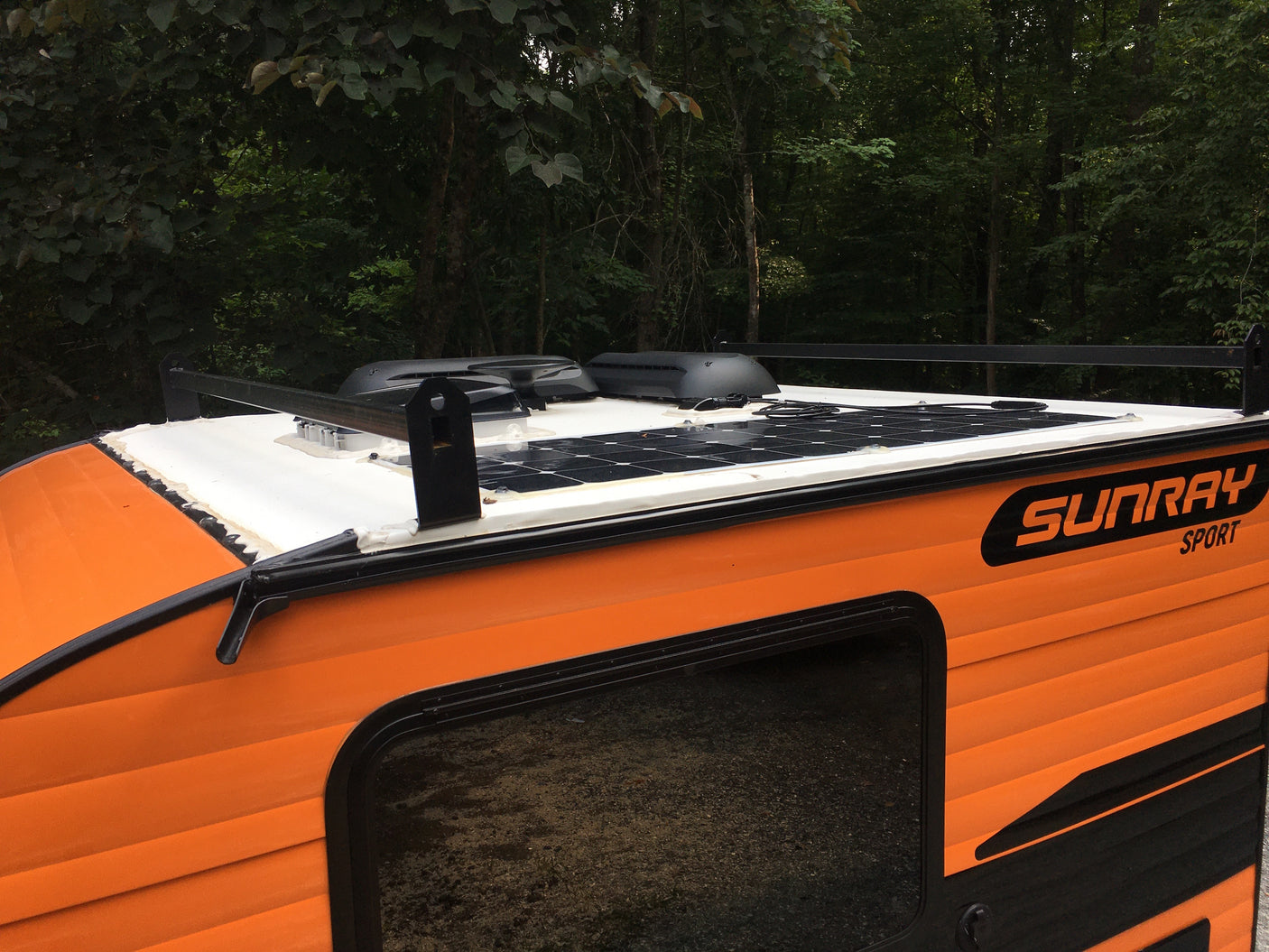 Close-up of an orange and black Sunray Sport rv rental, featuring roof-mounted solar panels & racks, amidst a forest.