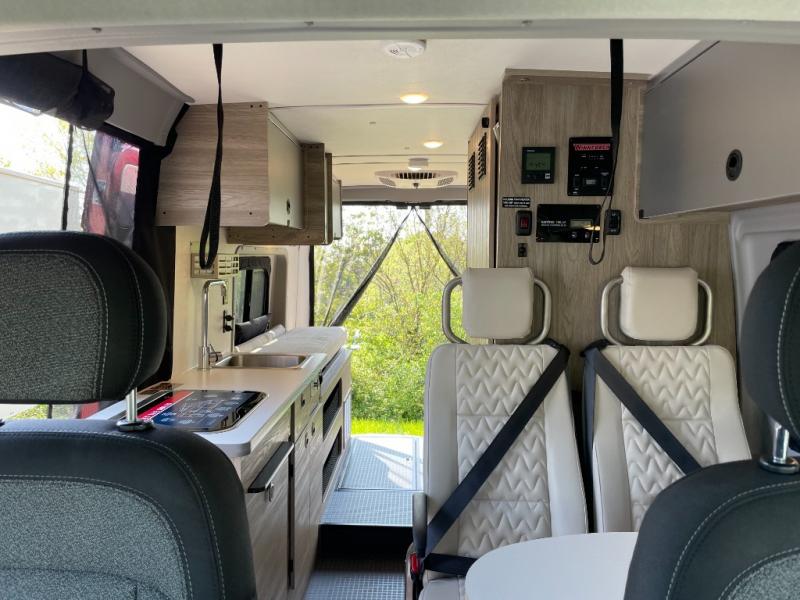 RV rental interior: kitchenette, two passenger seats, control panel, and rear exit to nature.