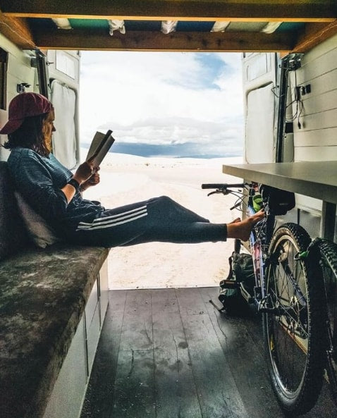 A woman reads in an RV rental, feet on her bike, gazing out at the vast desert. Perfect van life adventure!
