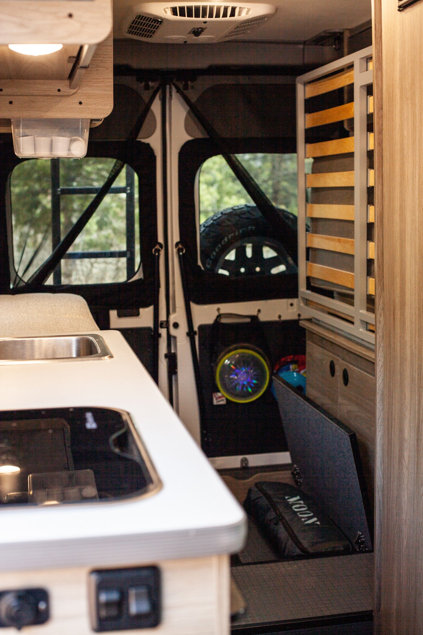 This RV rental interior features a compact kitchen with a sink and stovetop, integrated storage, and rear windows overlooking trees and a mounted spare tire.