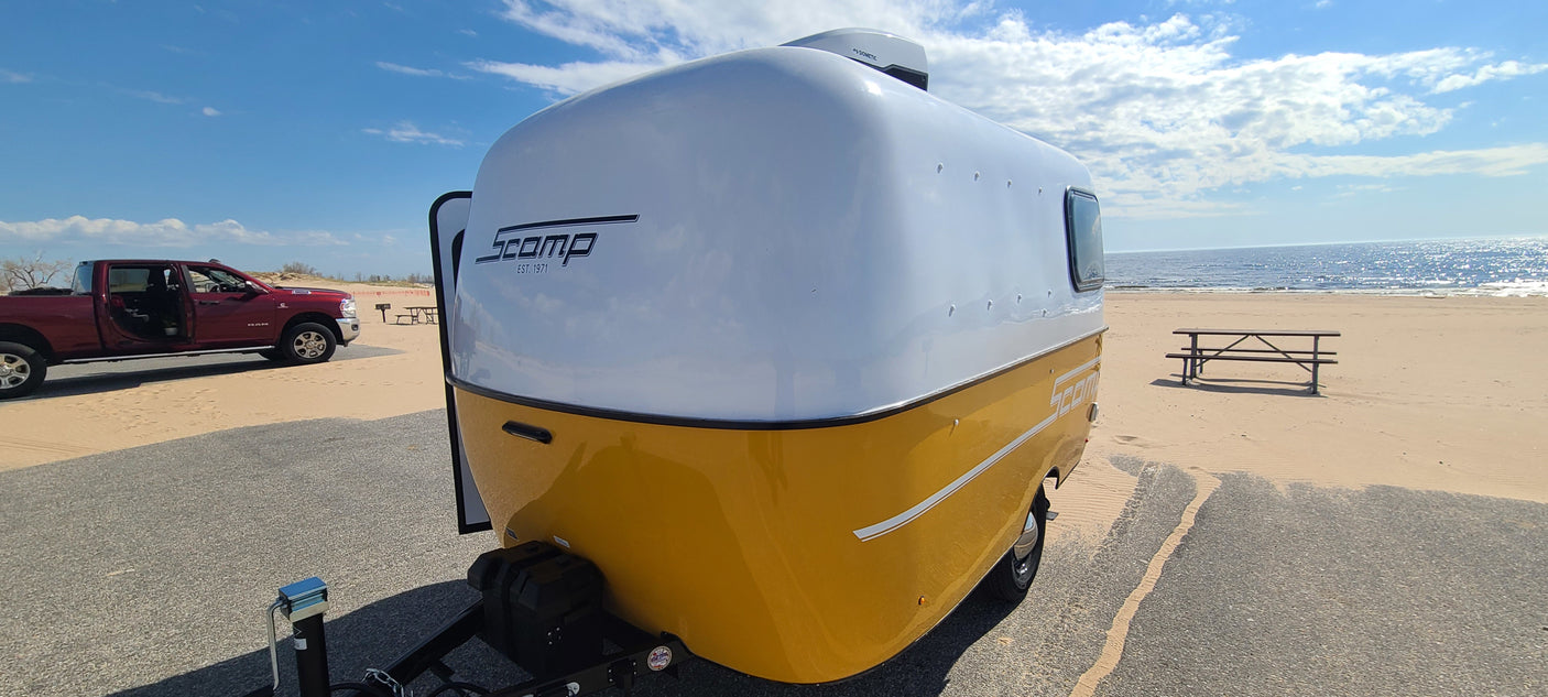 A white & yellow Scamp travel trailer (RV rental) and red truck are parked by a sandy beach with a picnic table & ocean.