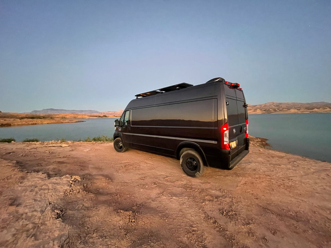 Black camper van, an ideal RV rental, parked lakeside on dirt. Distant mountains under a blue sky, perfect for outdoor adventure.