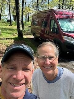 A smiling couple, a man in a cap and a woman with glasses, stand in front of their red RV rental at a wooded campsite.