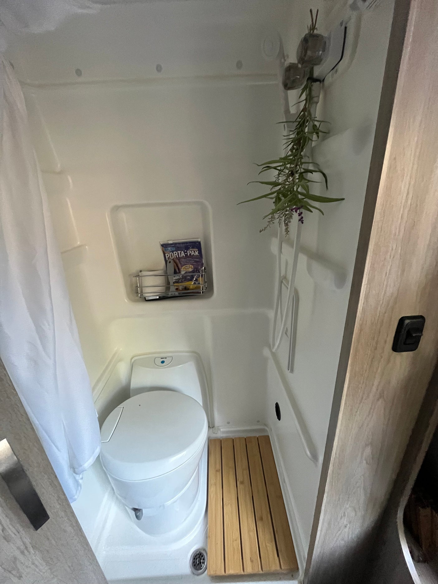 An RV rental's compact wet bath features a toilet, shower curtain, and a wooden floor mat, maximizing space.