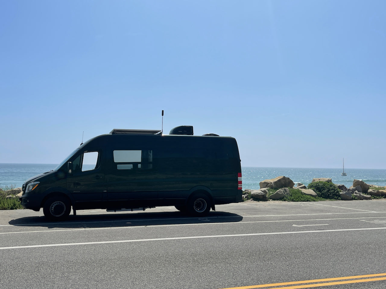 Dark green RV rental van parked by the sparkling blue ocean on a sunny day, with a distant sailboat. Scenic travel.