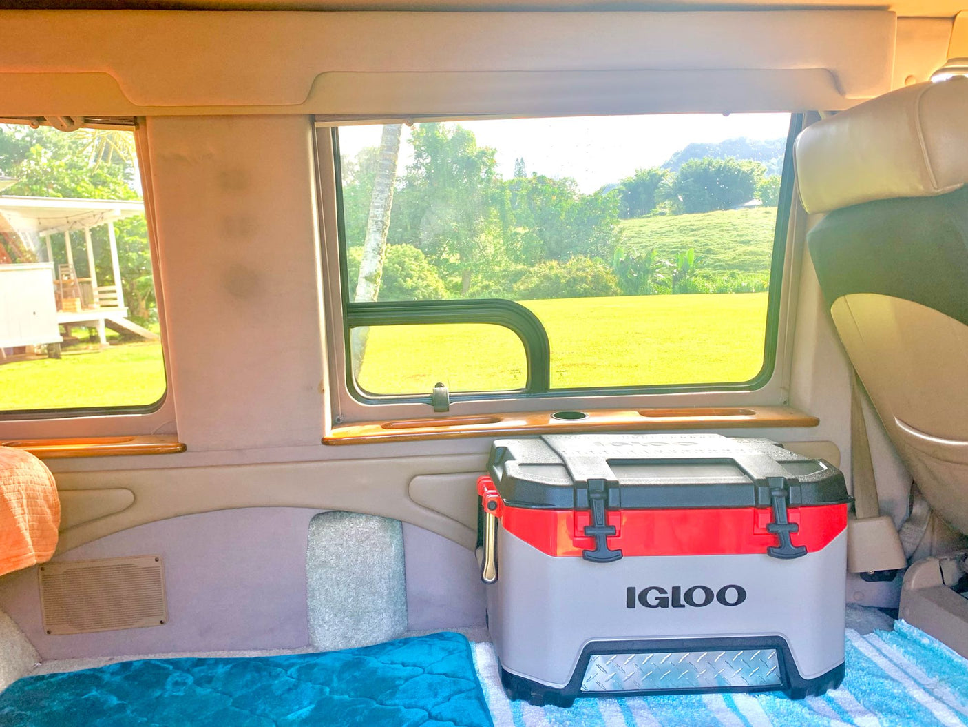 Inside an RV rental, a comfortable sleeping area with a blue blanket and a large Igloo cooler are visible, with lush green landscapes outside the windows.