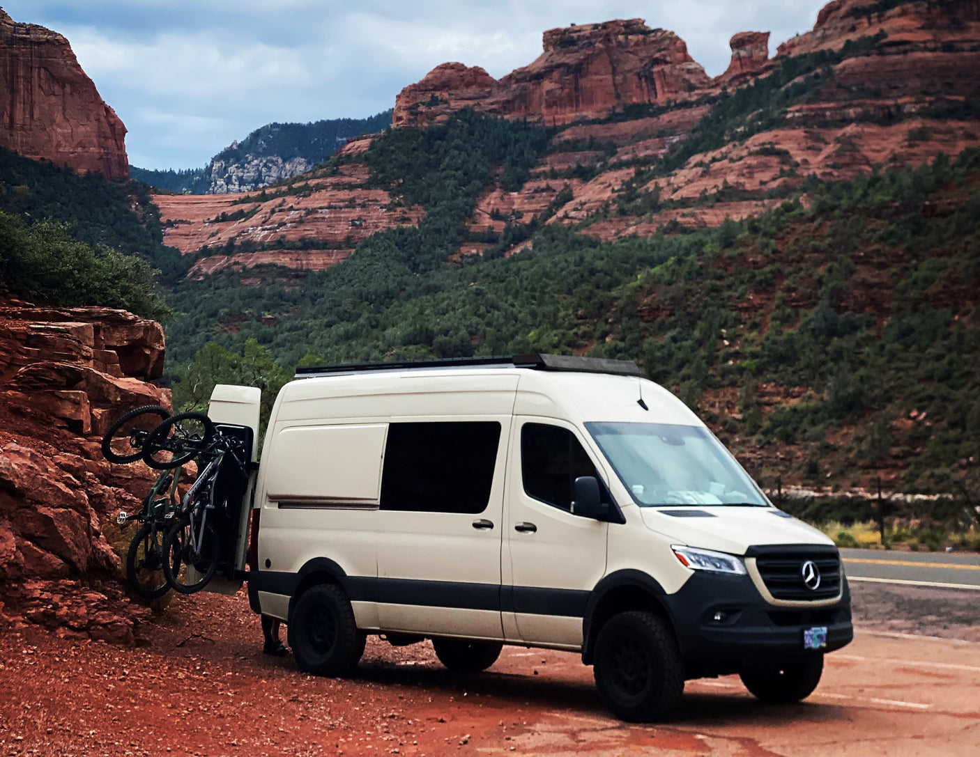 White RV rental camper van with bikes on a red dirt road, set against magnificent red rock mountains.