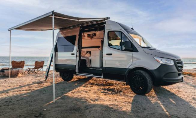 This bright RV rental camper van with an awning and chairs is set up for beach camping by the ocean.|Inside a modern RV rental: view of the camper van's custom interior with a versatile bunk bed system and orange cushions.|This white rv rental camper van