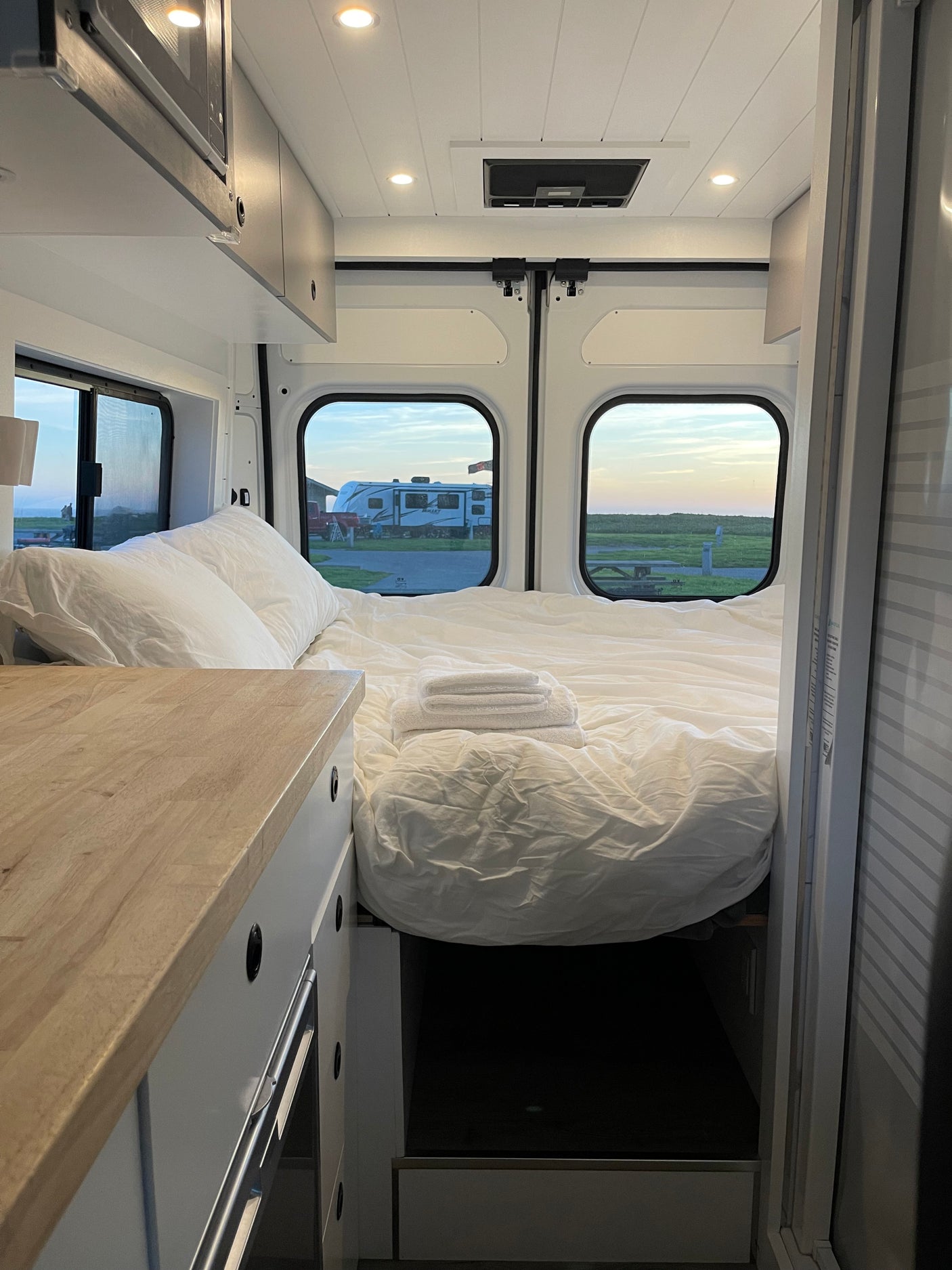 A comfortable and modern RV rental bed with white linens, towels, and picturesque outdoor views from the back windows.