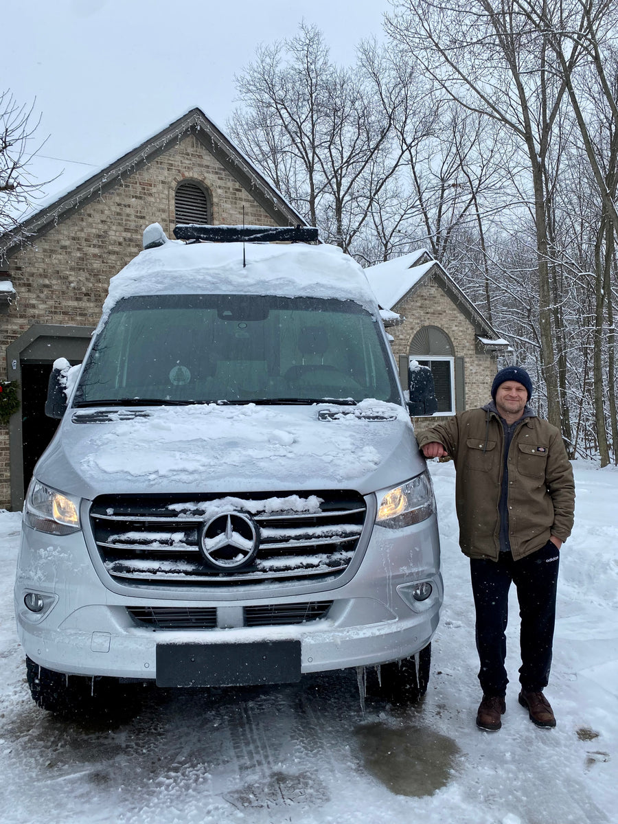 Man smiles next to his snow-covered Mercedes-Benz RV rental van, ready for winter travel adventures.