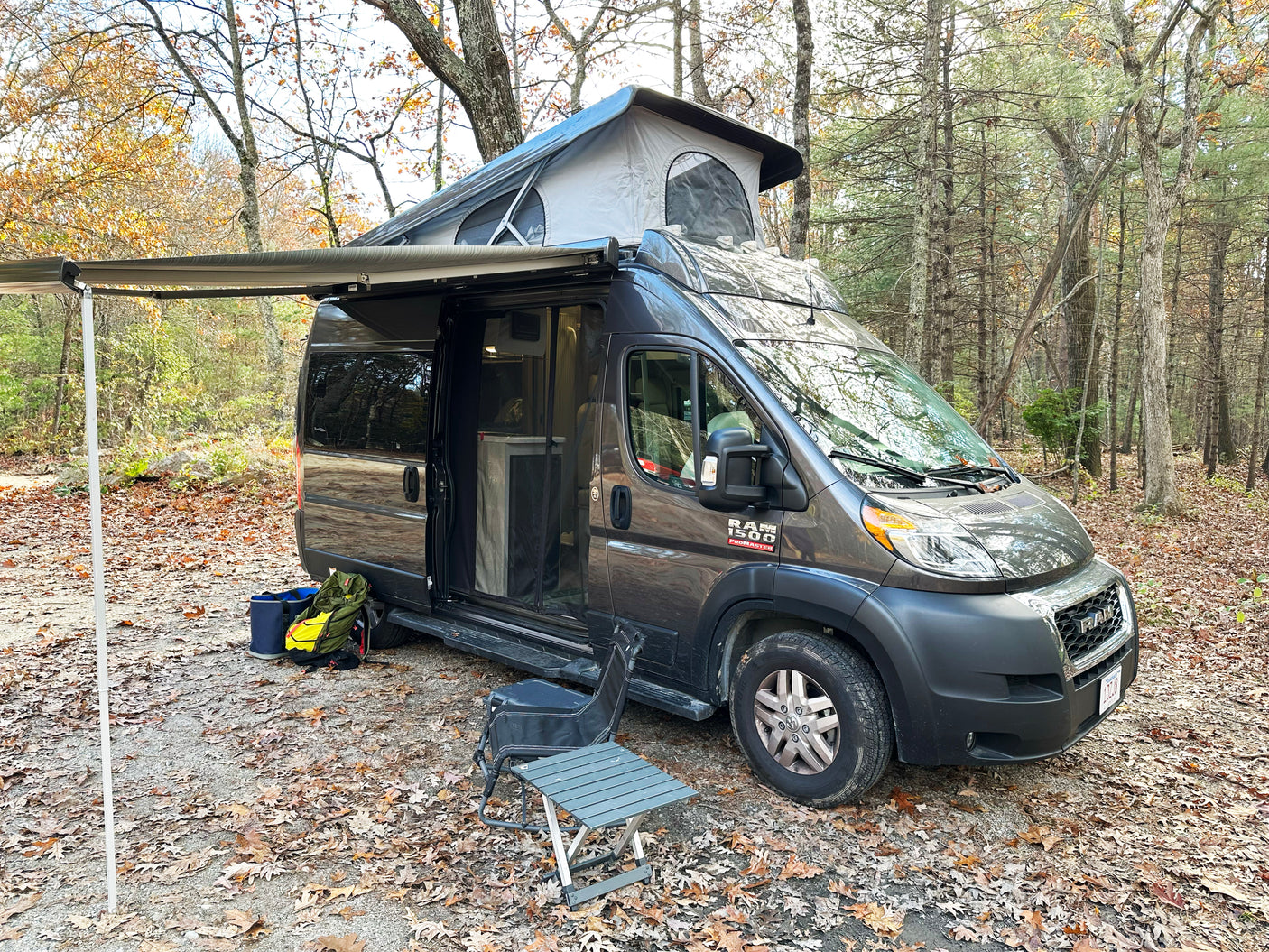 Grey Ram ProMaster RV rental with pop-top extended & awning out, parked in a scenic autumn forest campsite.