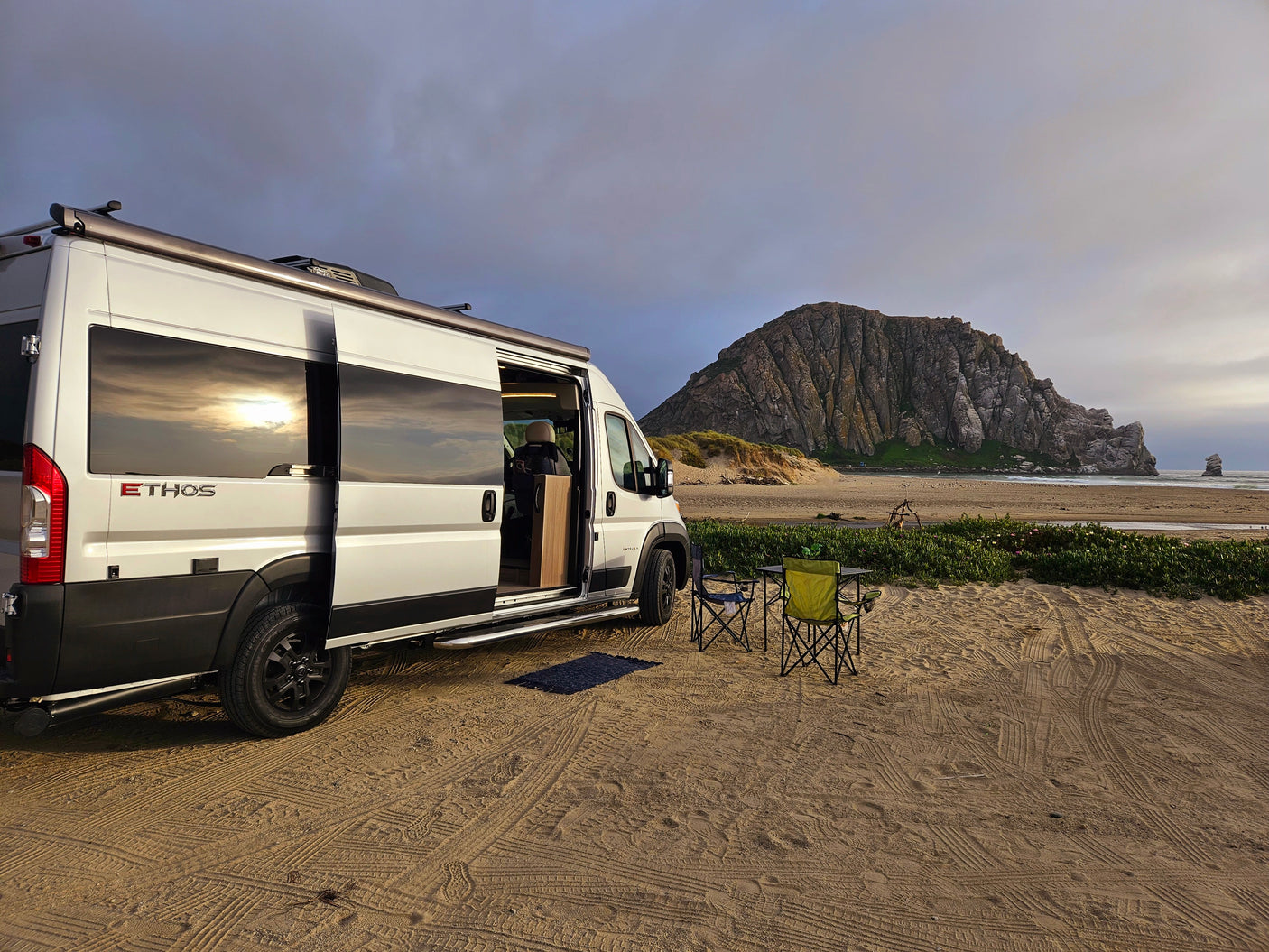 Scenic RV rental camping: a white van on a sandy beach with chairs, overlooking Morro Rock and the ocean.
