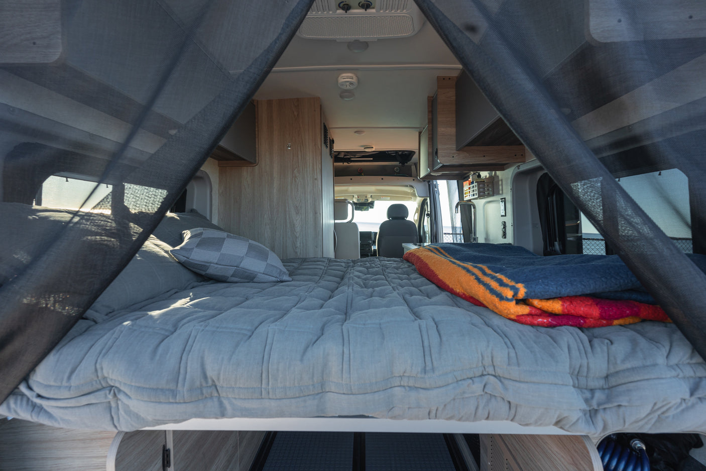 RV rental: Cozy camper van interior featuring a comfy grey bed with a colorful blanket, looking through netting to the front cab.