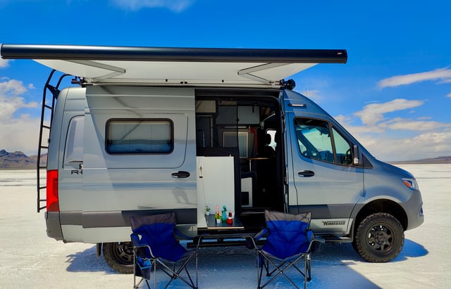 Custom gray camper van rv rental set up for adventure on a salt flat, with open door, extended awning, chairs & table.