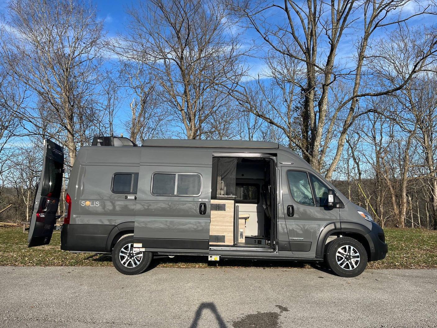 rv rental: A gray Winnebago Solis camper van with open doors, parked outdoors under bare trees on a sunny day.