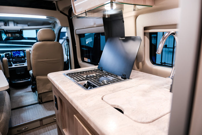 RV rental: Inside a camper van, showing a compact kitchen with a stove and sink in the foreground and the driver's cabin beyond.
