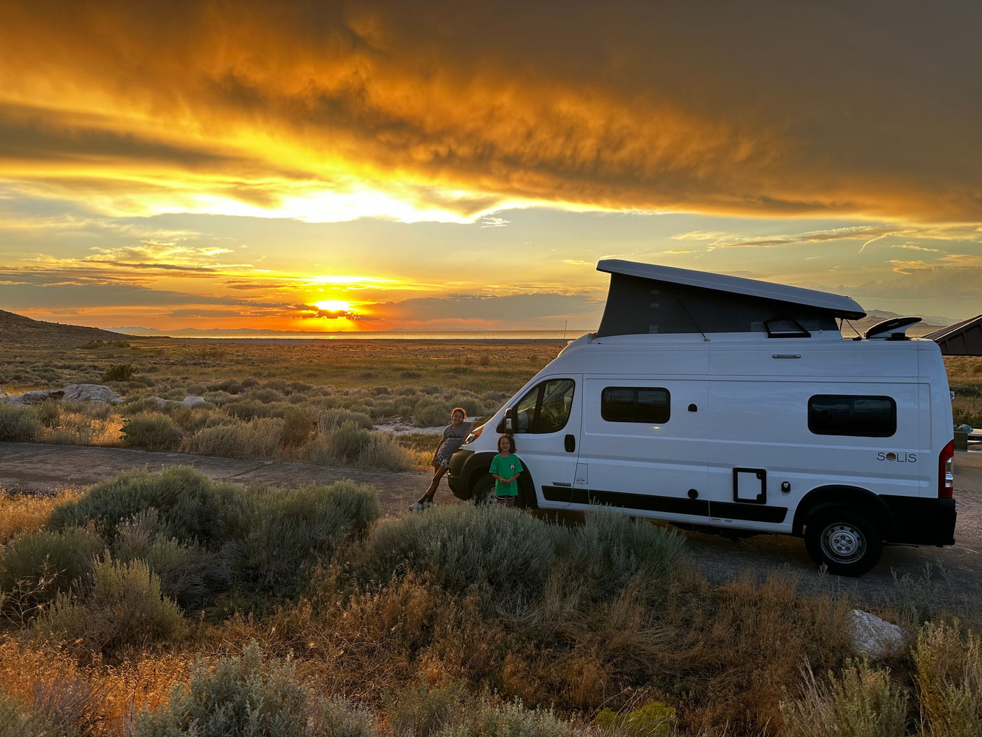 Two people enjoy a stunning sunset beside their RV rental, set against a vast, open landscape, perfect for an outdoor adventure.